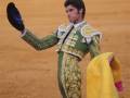 Un torero en traje tradicional de color verde y dorado, con un sombrero azul y una capa amarilla, en una plaza de toros.