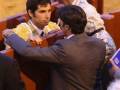 Un torero en traje de terciopelo y corbata, con un chaleco amarillo, se dirige a una persona en traje negro. La imagen muestra un ambiente de corrida de toros, con asistentes y público en el fondo.