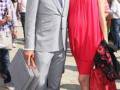 Un hombre en traje gris y corbata azul, sosteniendo una maleta de viaje, camina junto a una mujer en vestido rojo y zapatos rojos. Ambos parecen estar en un evento formal, con otros asistentes de fondo.