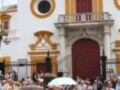 Fotografía de una celebración en una plaza con personas vestidas de manera tradicional, sosteniendo paraguas y sentadas en bancos. El edificio en el fondo es de estilo colonial, con detalles arquitectónicos y una puerta principal roja.