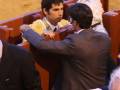 Un hombre en traje negro se inclina hacia otro hombre en traje, ambos están sentados en un banco de madera. El hombre en el traje negro parece estar hablando o explicando algo al otro, mientras que el hombre en traje está escuchando atentamente. En el fondo, se pueden ver otros asistentes sentados en bancos similares, algunos de ellos con sombreros. La escena parece ser una reunión o conferencia formal en un entorno de arena o estadio.