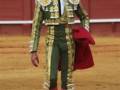 Un torero en traje tradicional de oro y verde, con guantes rojos y calcetines rosados, en una plaza de toros.