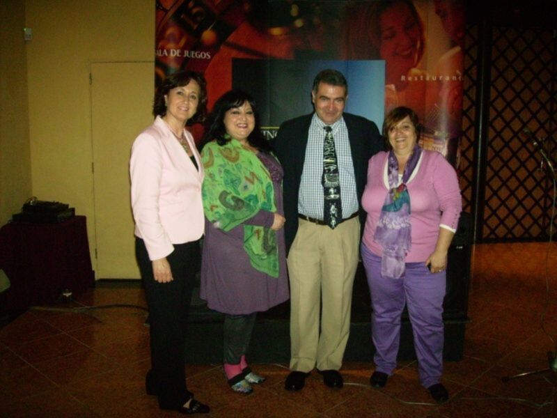 Una mujer en una chaqueta rosa, otra con un vestido verde y una falda morada, un hombre en una camisa blanca y una corbata negra, y otra mujer con una bufanda morada. Todos están en un ambiente festivo con carteles de juegos al fondo.