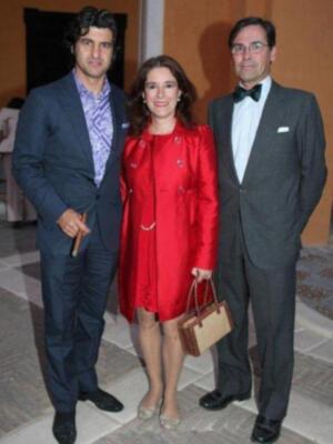 Tres personas posan juntas en un evento formal. La persona en el centro lleva una chaqueta roja y sostiene una cartera beige. A la izquierda, un hombre viste un traje azul con camisa a cuadros. A la derecha, otro hombre está vestido con un traje negro y corbata verde. El fondo muestra una alfombra y parte de otro individuo en traje blanco.