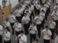 Una banda musical en uniforme blanco y negro, tocando instrumentos de viento, posiblemente una trompeta o tuba. La imagen parece ser tomada desde arriba, mostrando a los músicos en una fila.