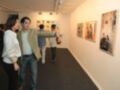 Un hombre y una mujer observan con interés una exposición de arte en un museo. La pared está llena de cuadros y fotos, mientras que el suelo es de color gris oscuro. La iluminación del lugar es adecuada para la exposición, y las personas parecen estar disfrutando de la experiencia.