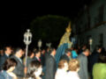 Una procesión nocturna en una calle con gente vestida formalmente. La imagen muestra a una figura religiosa vestida de azul y dorado, seguida por un grupo de personas. Hay faroles y una placa indicativa en el fondo.