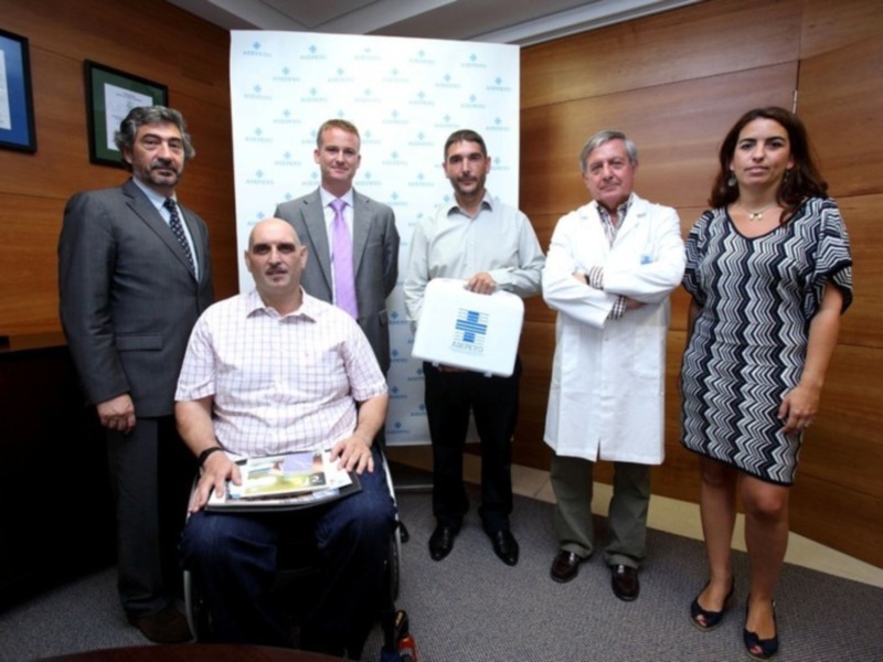 Equipo médico y personalidades en una reunión formal, con un hombre en silla de ruedas y una persona en vestido negro, en un entorno profesional con un fondo de logotipo.