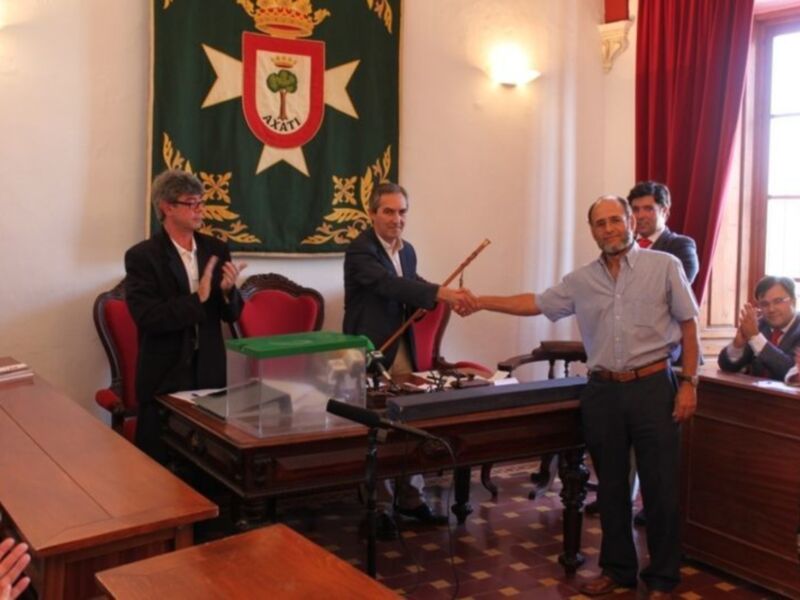 Un hombre en una sala de conferencias sostiene un bastón mientras otros asistentes aplauden. En el fondo, se puede ver una bandera con un escudo y un árbol. La sala tiene una mesa con una urna de votación y microfonios en el centro.