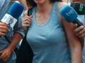 Una mujer con gafas de sol y una camiseta azul está siendo entrevistada por dos micrófonos. Detrás de ella, hay personas con pantalones cortos y sandalias, posiblemente en un evento o protesta.