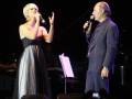 Dos personas cantando en un escenario, una con un vestido largo y la otra con una camisa negra.