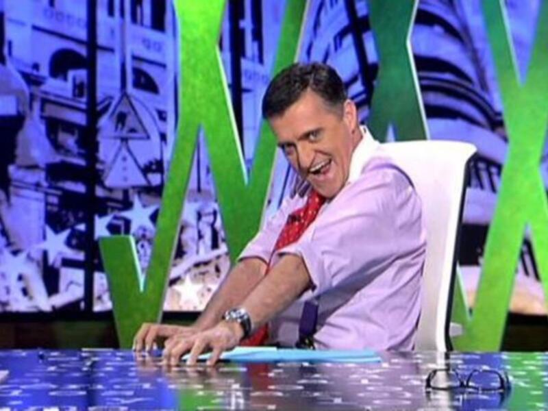 Un presentador de televisión en un set de noticias, sonriendo y trabajando en el escritorio.