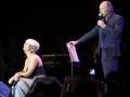 Una mujer con cabello corto y blanco está sentada en un piano, mientras un hombre mayor con una camisa azul y pantalones negros se encuentra frente a ella, sosteniendo un micrófono. La escena parece ser una actuación musical en vivo.
