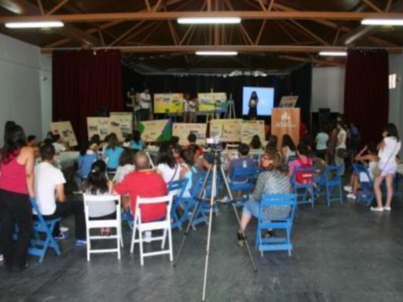 Un auditorio lleno de personas sentadas en sillas, mirando a un escenario con un panel de presentaciones.