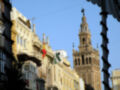 Imagen de una ciudad con edificios históricos y la Giralda, una famosa torre en el horizonte. Banderas ondean en las fachadas, y el cielo es azul claro.