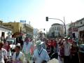 Desfile en una ciudad con edificios amarillos y azules, gente vestida de colores, banderas y carteles publicitarios.