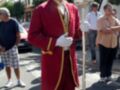 Un hombre vestido con un traje rojo y dorado, posiblemente para una celebración o evento formal, camina por la calle mientras sostiene un bastón. A su lado, otros individuos observan el evento. La imagen muestra una calle con edificios y un cartel de止止。
