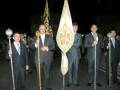 Grupo de personas en trajes formales, posiblemente en una procesión religiosa o ceremonial nocturno. Algunos sostienen bastones decorativos y una bandera con emblemas. La escena parece ser nocturna, iluminada por luces de fondo.