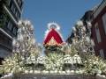 Fotografía de una procesión religiosa con una imagen venerada en el centro, rodeada de flores blancas y flanqueada por dos estatuas con luces. El cielo es azul claro y las edificaciones alrededor sugieren una ciudad histórica.