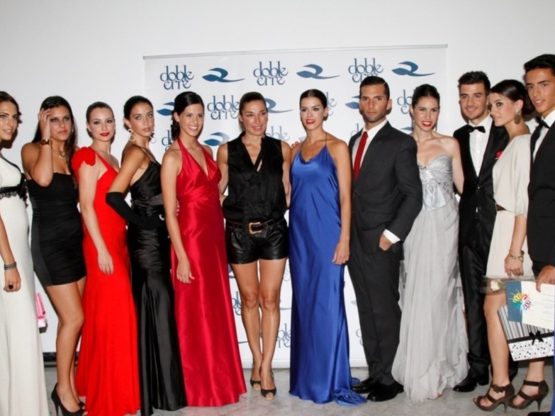 Una fila de personas vestidas con elegantes vestidos de noche, incluyendo un vestido rojo, un vestido negro, un vestido azul y un vestido blanco, posando frente a un fondo con la marca 'dbbe' repetida.