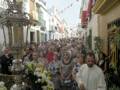 Una multitud en procesión por una calle, con banderas y luces colgadas.