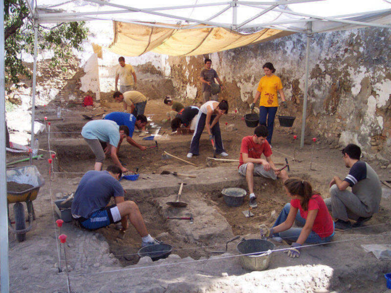 Un grupo de personas trabajando en una excavación arqueológica bajo un toldo, con herramientas y materiales de excavación dispersos alrededor.