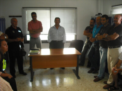 Un grupo de personas se encuentra en una sala con una mesa central. Algunos están de pie, mientras que otros tienen las manos cruzadas sobre los hombros. La sala tiene ventanas con cortinas y una persona en la esquina izquierda parece estar hablando.