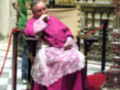 El papa Francisco, vestido con un hábito rojo y rosa, se sienta en una silla de madera con un tapizado rojo. A su lado, un hombre vestido verde observa la escena. La imagen se encuentra en una iglesia con detalles dorados y una alfombra roja.