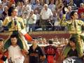 Imagen de una plaza de toros con dos matadores en trajes tradicionales, uno llevando un toro muerto. El público observa el evento.