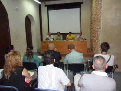 Una sala de conferencias con un grupo de personas sentadas en sillas frente a una mesa donde están los presentadores. La pared tiene un gran proyector en la parte superior y una columna de piedra en el lado derecho.
