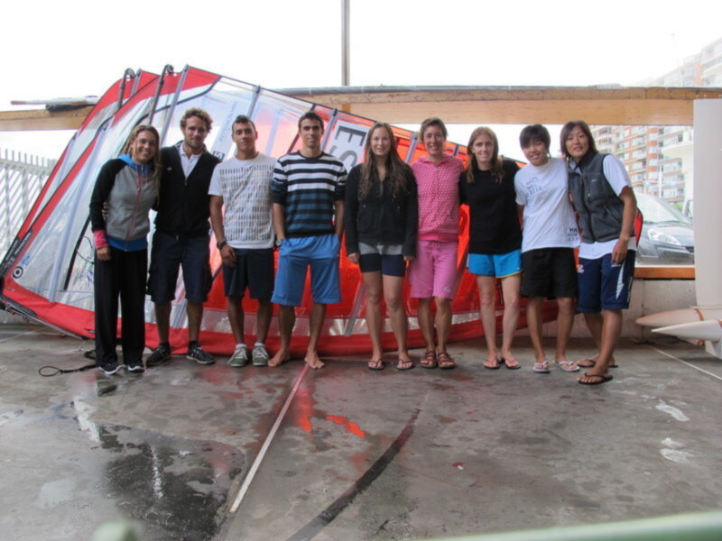 Grupo de personas posando frente a una carpa de windsurf en una playa con edificios en el fondo.