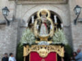 Fotografía de una procesión religiosa con una imagen de la Virgen en un carro adornado con flores y velas, rodeada de personas observando la celebración.