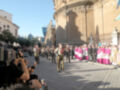 Desfile militar en una ciudad histórica con soldados armados y uniformes militares, gente en trajes tradicionales de pie junto a un edificio monumental.