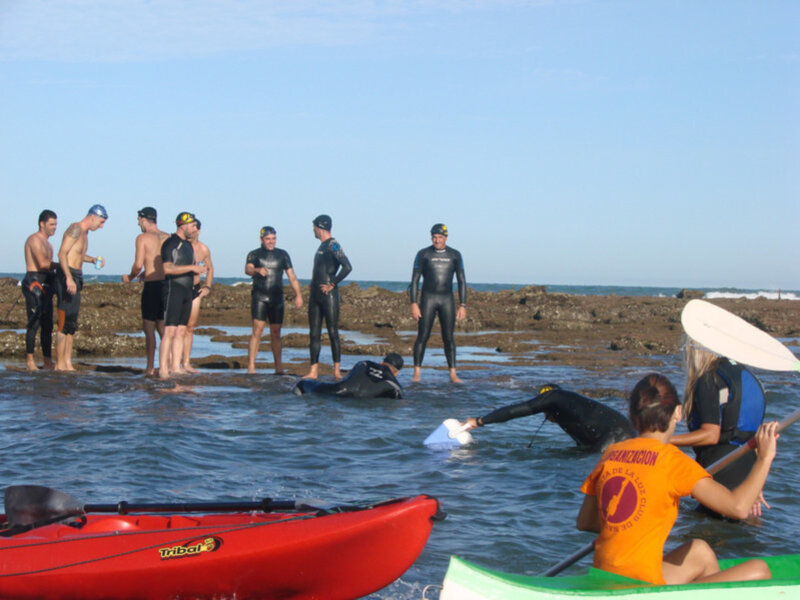 Un grupo de personas en trajes de neopreno y ropa de surf en la orilla del mar, preparándose para la actividad de surf.