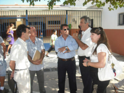 Un grupo de personas conversando al aire libre frente a una escuela. Un hombre en el centro tiene un libro y gafas, mientras que una mujer a su derecha lleva una bolsa blanca. La escuela tiene ventanas y un azulejo en la pared.