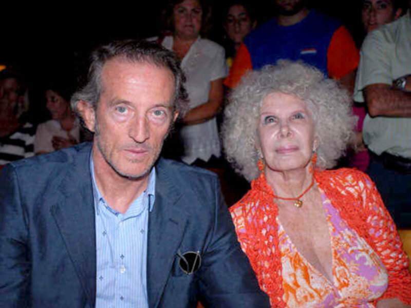 Dos personas, una mujer con cabello blanco y una persona masculina, están sentados en un evento público. La mujer lleva una chaqueta naranja y una camisa con estampado, mientras que el hombre está vestido con un traje azul. La escena parece ser una reunión o conferencia, con otros asistentes en el fondo.