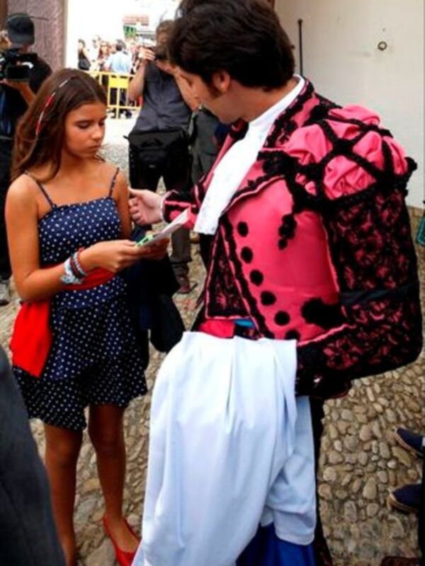 Un hombre en traje de carnaval rojo y negro con un chaleco blanco está firmemente firmando una tarjeta de visita para una mujer en un vestido azul con lunares. Ambos están en una calle de piedra, con otros individuos y edificios al fondo.