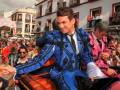 Un hombre en traje de torero azul y negro se sienta en un carro durante una celebración festiva. Detrás de él, personas vestidas con trajes tradicionales y una mujer en un traje rosa están observando la celebración. La imagen se desarrolla frente a una casa blanca con ventanas y balcones, donde otros espectadores observan la celebración.