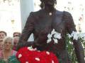 La estatua de un personaje histórico, con un sombrero elaborado y una corona floral roja en la cabeza, es el centro de atención. Detrás de ella, se pueden ver personas observando la escultura, y hay un fondo con elementos decorativos que acentúan el ambiente festivo.