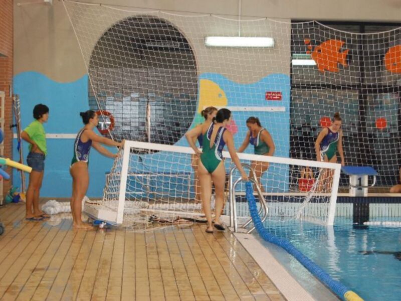 Niñas jugando a voleibol de agua en una piscina interior con una red grande y varios jugadores en trajes de baño.