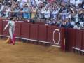 Un torero en traje tradicional se prepara para entrar en la plaza de toros. La audiencia, compuesta por personas vestidas con ropa formal y accesorios típicos de la cultura taurina, observa atentamente el evento. La arena está rodeada por una cerca roja con un escudo blanco en su centro, típico de las plazas de toros españolas. La arena está cubierta con tierra, típica del ambiente de la tauromaquia.