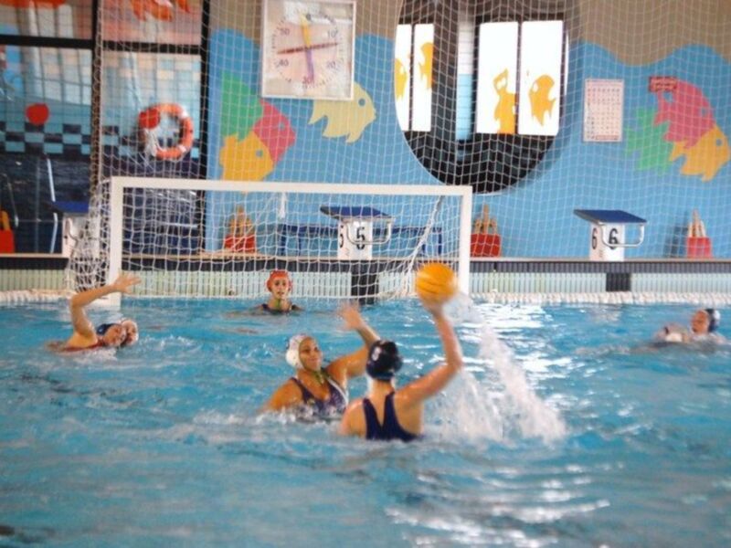Un equipo de waterpolo en acción, con una jugadora lanzando el balón hacia la portería mientras otros jugadores observan.