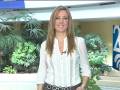 La presentadora sonríe mientras está en una estación de noticias, con un fondo de plantas y un logotipo visible.