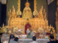 Fotografía de una iglesia con un altar dorado y una imagen religiosa en el centro. Personas rezando frente al altar, con velas encendidas y flores decorativas. El ambiente es reverente y solemnemente religioso.