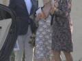Una mujer anciana con vestido de encaje blanco y zapatos de tacón negro, abraza a una mujer joven con vestido floral oscuro. Un hombre de traje azul y gafas de sol los observa desde el lado. Detrás, un coche negro y una casa con puerta de madera.