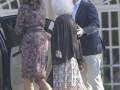 Una mujer con vestido floral y zapatos de tacón negro se acerca a un hombre en chaqueta azul y pantalones beige, mientras una mujer con vestido de lunares y abrigo negro observa. El fondo muestra un edificio blanco con ventanas grandes.