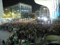 Una multitud de personas reunidas en una plaza pública, con edificios modernos al fondo y luces brillantes iluminando la noche.