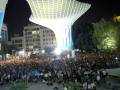 Una multitud de personas disfruta de un concierto en la noche bajo una estructura futurista iluminada, con árboles y edificios alrededor.
