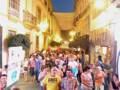 Una multitud camina por una calle de noche iluminada, con edificios históricos a ambos lados. La gente parece disfrutar de una celebración o evento, con algunos portando pancartas y camisetas. La atmósfera es festiva, con luces brillantes y un cielo nocturno claro.