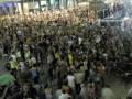 Una multitud en un evento público, con personas de todas las edades y vestimentas variadas. El ambiente parece ser animado, con algunos participantes levantando las manos y otros posiblemente tomando fotos o videos. La escena ocurre en un lugar con estructuras de acero y grandes pantallas, posiblemente una plaza o estación de transporte.